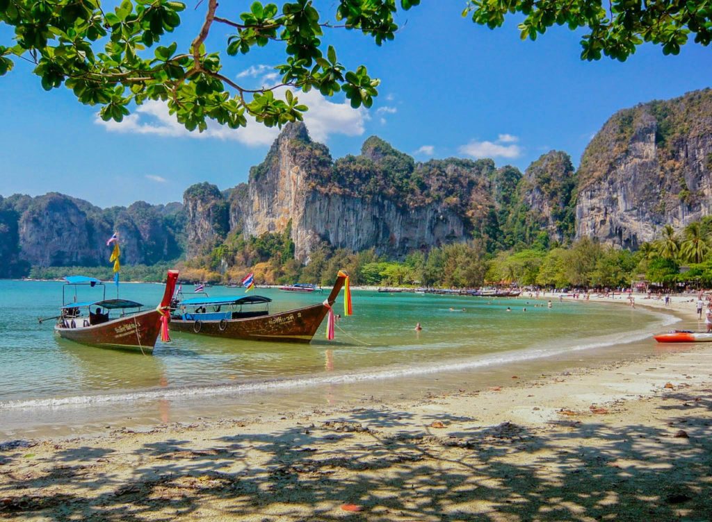 A Thailand beach with colorful boats in the ocean and mountains in the background.