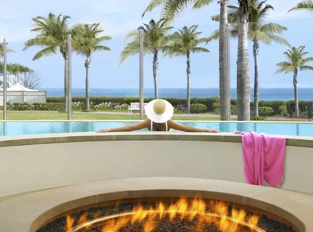 Woman lounging in a pool overlooking the ocean with a firepit behind her.