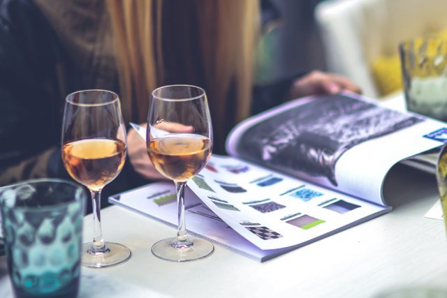 Person reading a magazine at a table with two wine glasses.