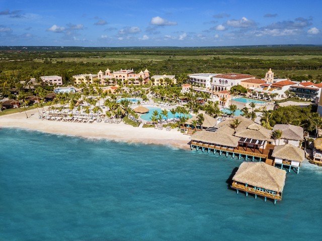 Aerial view of Sanctuary Cap Cana resort in the Dominican Republic. There are while buildings surrounding multiple pools and an overwater hut with a thatched roof in the ocean.