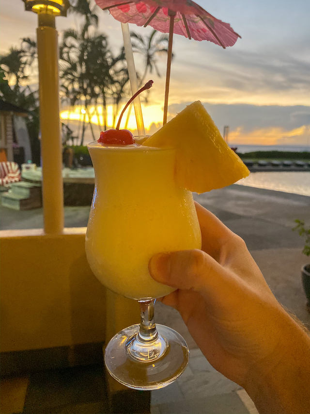 Person holding a yellow cocktail with a pink umbrella, pineapple wedge, and a cherry.