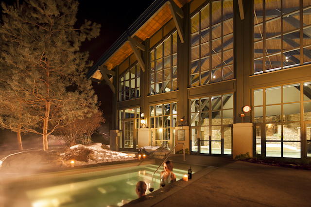 A couple relaxes in an outdoor pool at night with wine just outside a full glass enclosed building.