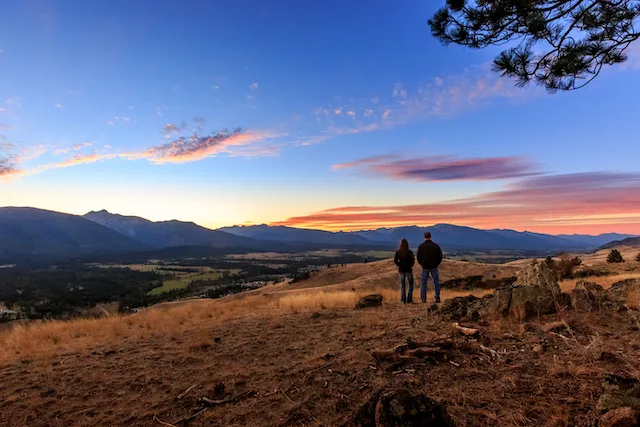 A couple standing in an open space overlooking a sunset, rolling hills, and distant mountains.