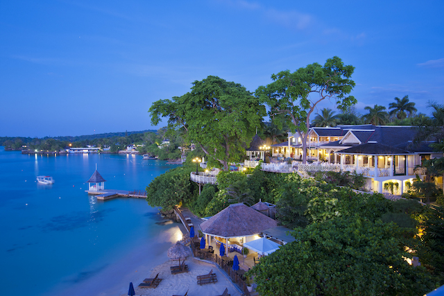 Scenic view of a coastal resort at twilight with buildings, trees, a gazebo by the water, and a sandy beach with lounge chairs and umbrellas.