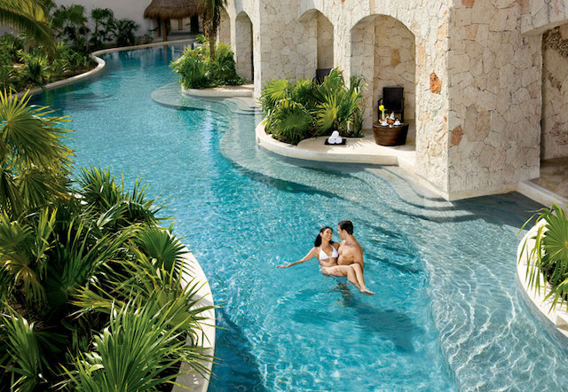 A man holds a woman while floating in a clear blue pool outside a resort.