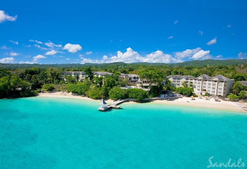 Clear blue ocean water in the foreground with a sandy beach and lush vegetation surrounding large cream hotel buildings.