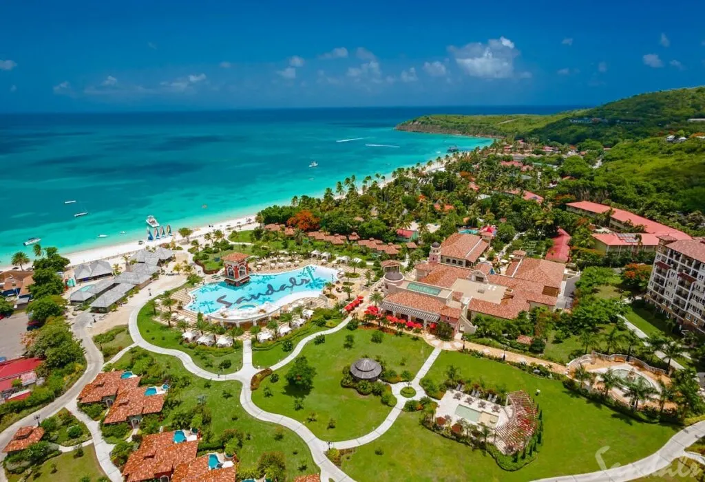 An aerial view of a Sandals resort with a beach, ocean, and hills in the background.