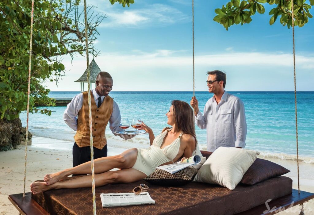 Sandals Royal Plantation resort - a woman on a beach front hammock is served a drink by a butler, romantic partner stands behind her.