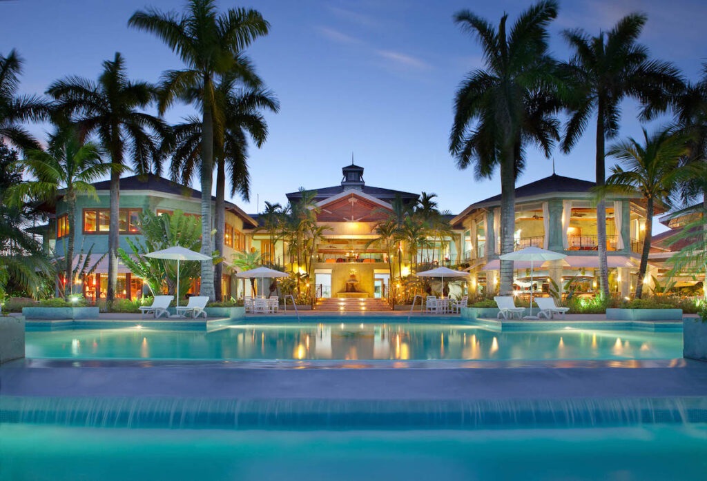 A front view of a well-lit resort with a pool, palm trees, and lounge chairs.