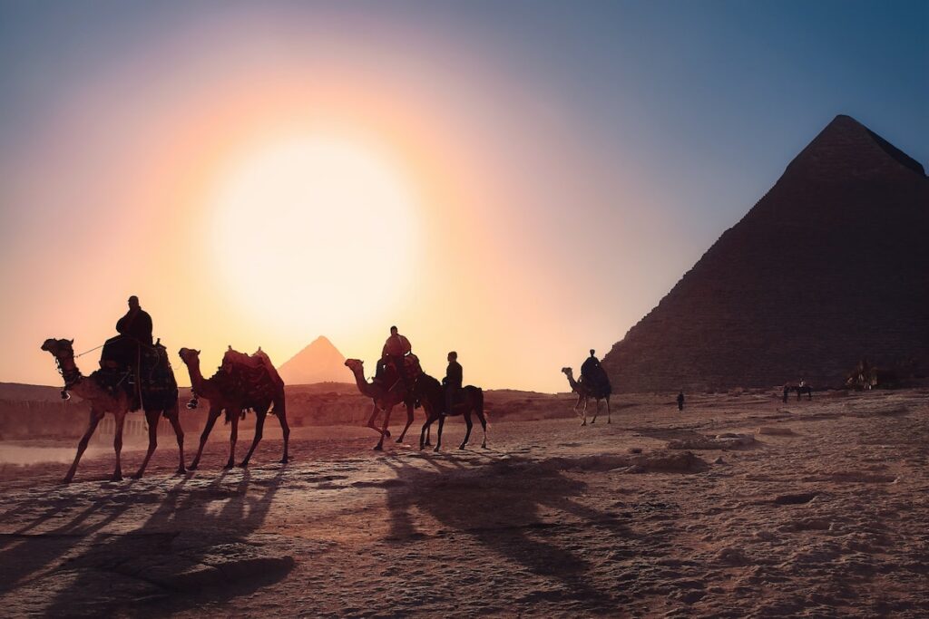Silhouette of men riding camels in the shadow of pyramids.