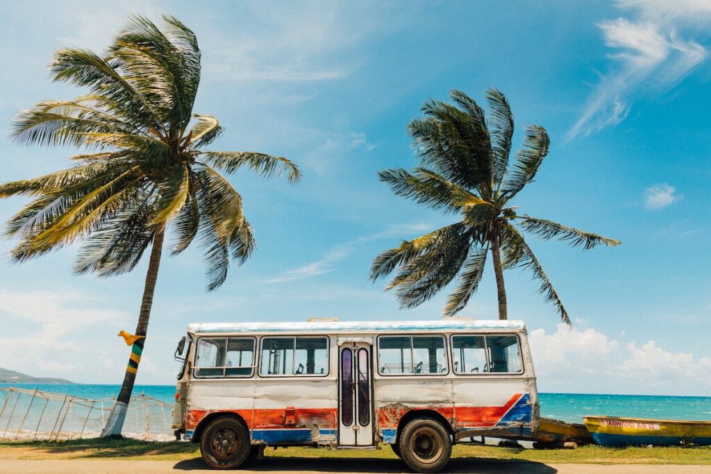 An older white van on a beach with palm trees and the ocean in the background.