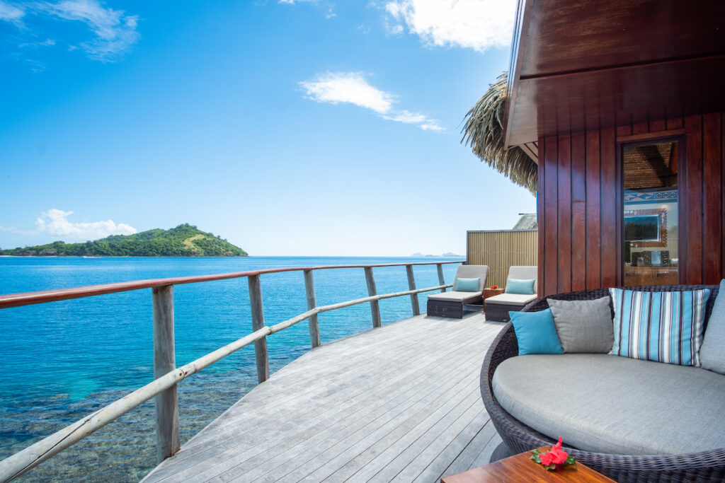 Deck of an overwater bungalow overlooking ocean and a treed island in the distance.
