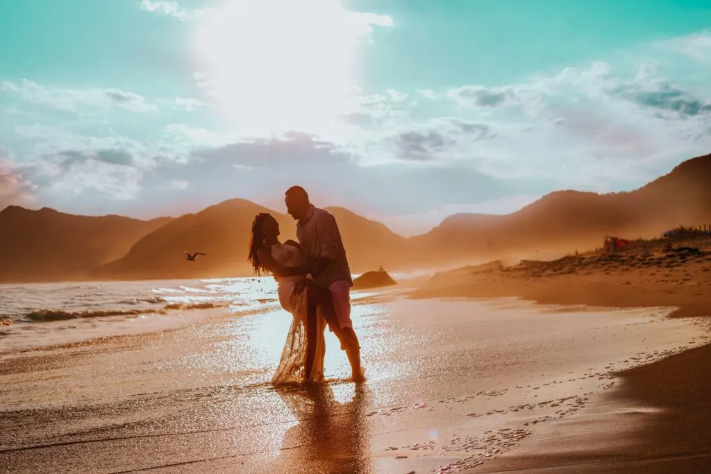 Couple dancing on a sandy beach with hills and mountains in the background.