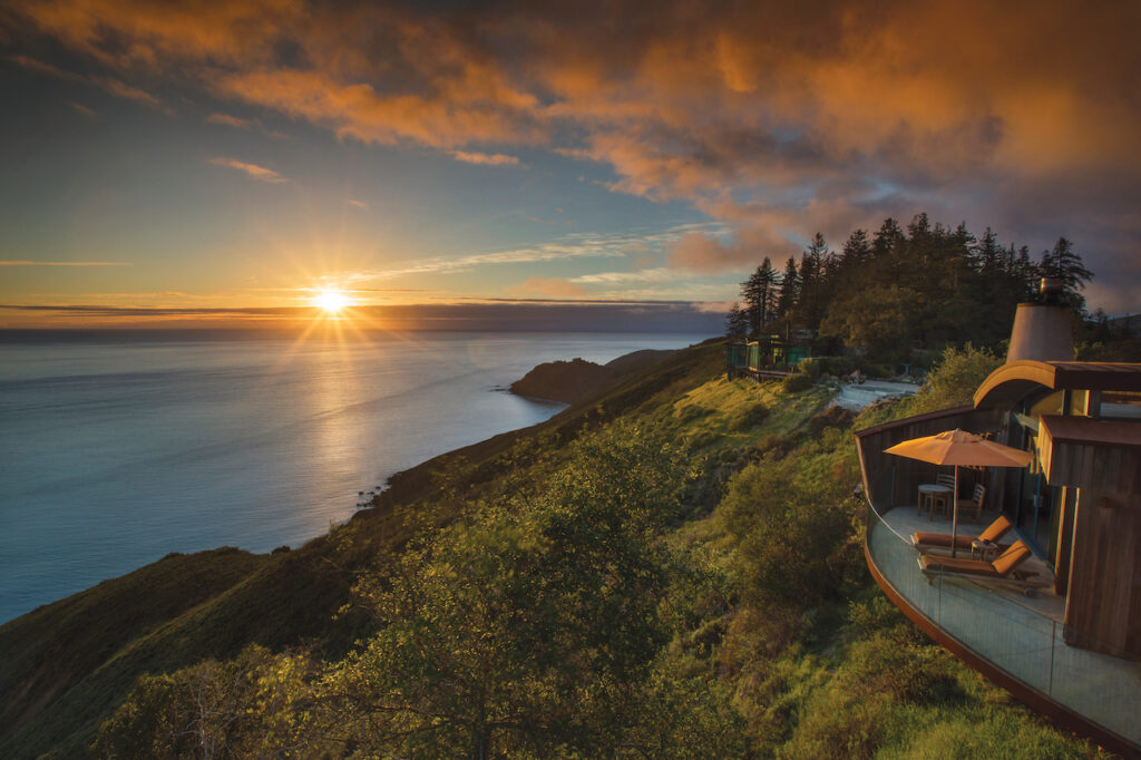 The back deck of a home with two lounge chairs overlooking a green cliff and a large body of water at sunset.