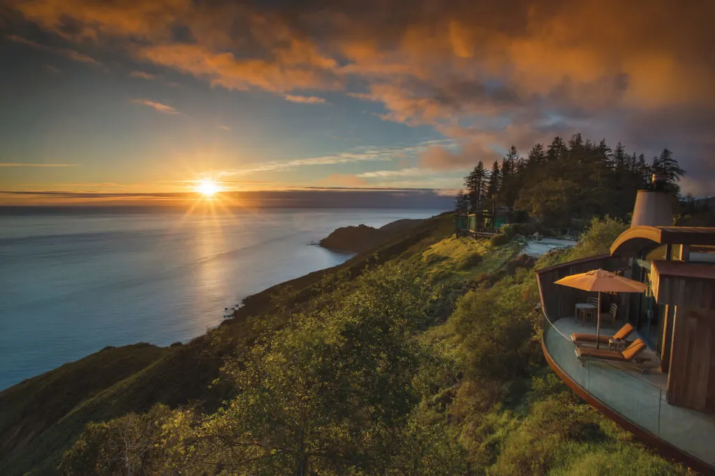 The back deck of a home with two lounge chairs overlooking a green cliff and a large body of water at sunset.