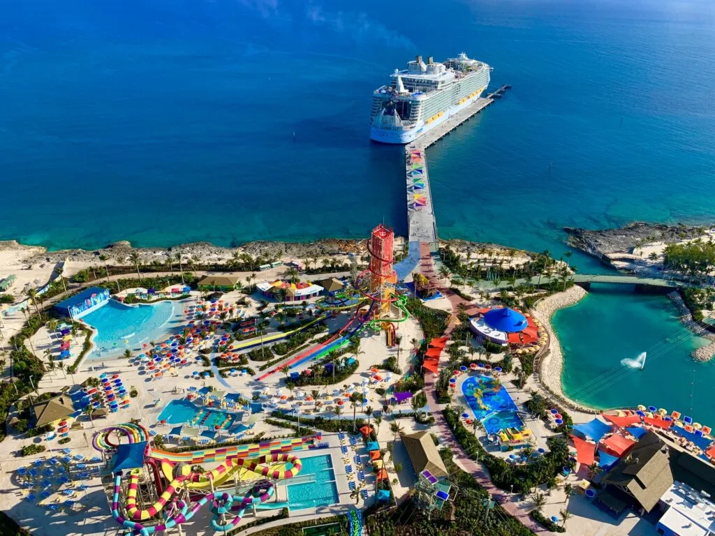 Arial view of a docked cruise ship in port filled with slides, pools, and colorful umbrellas over lounge chairs.