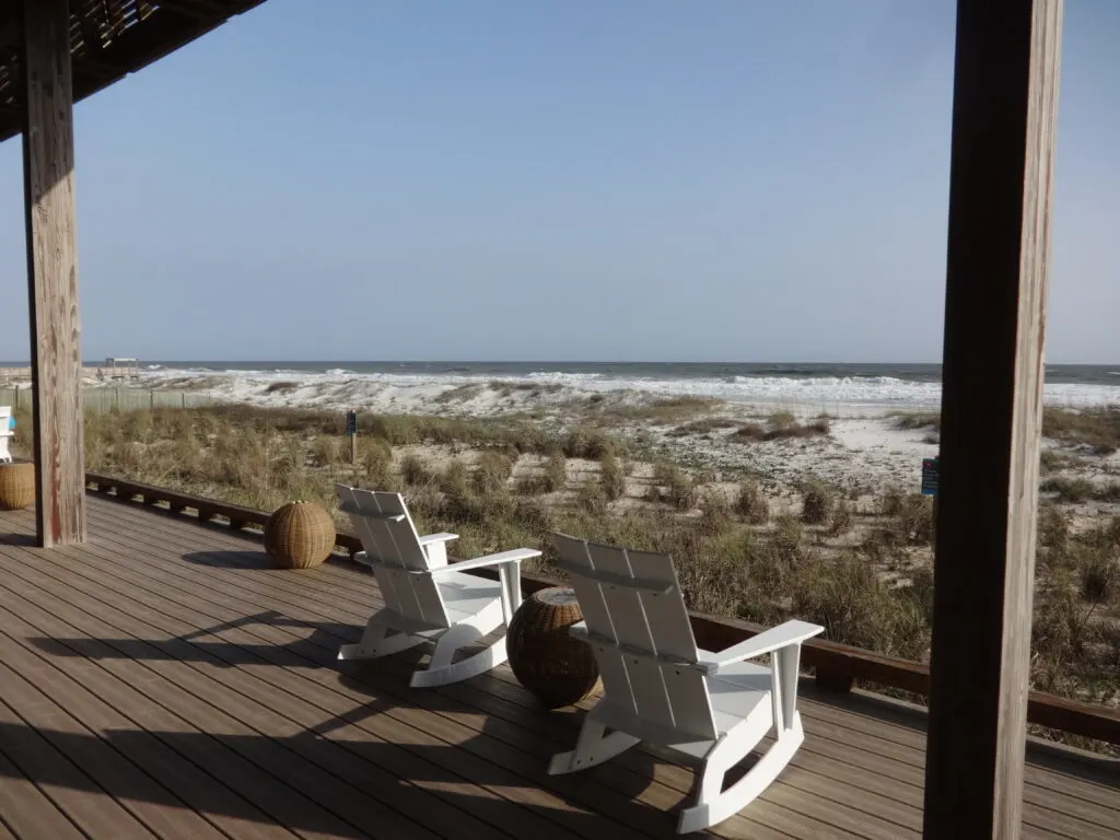 Two white rocking chairs sitting on a deck overlooking the sand and ocean.