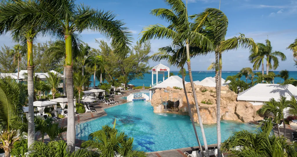 Outdoor swimming pool surrounded by palm trees, lounge chairs, and cabanas, with a rock formation, white canopies, and the ocean visible in the background under a blue sky.