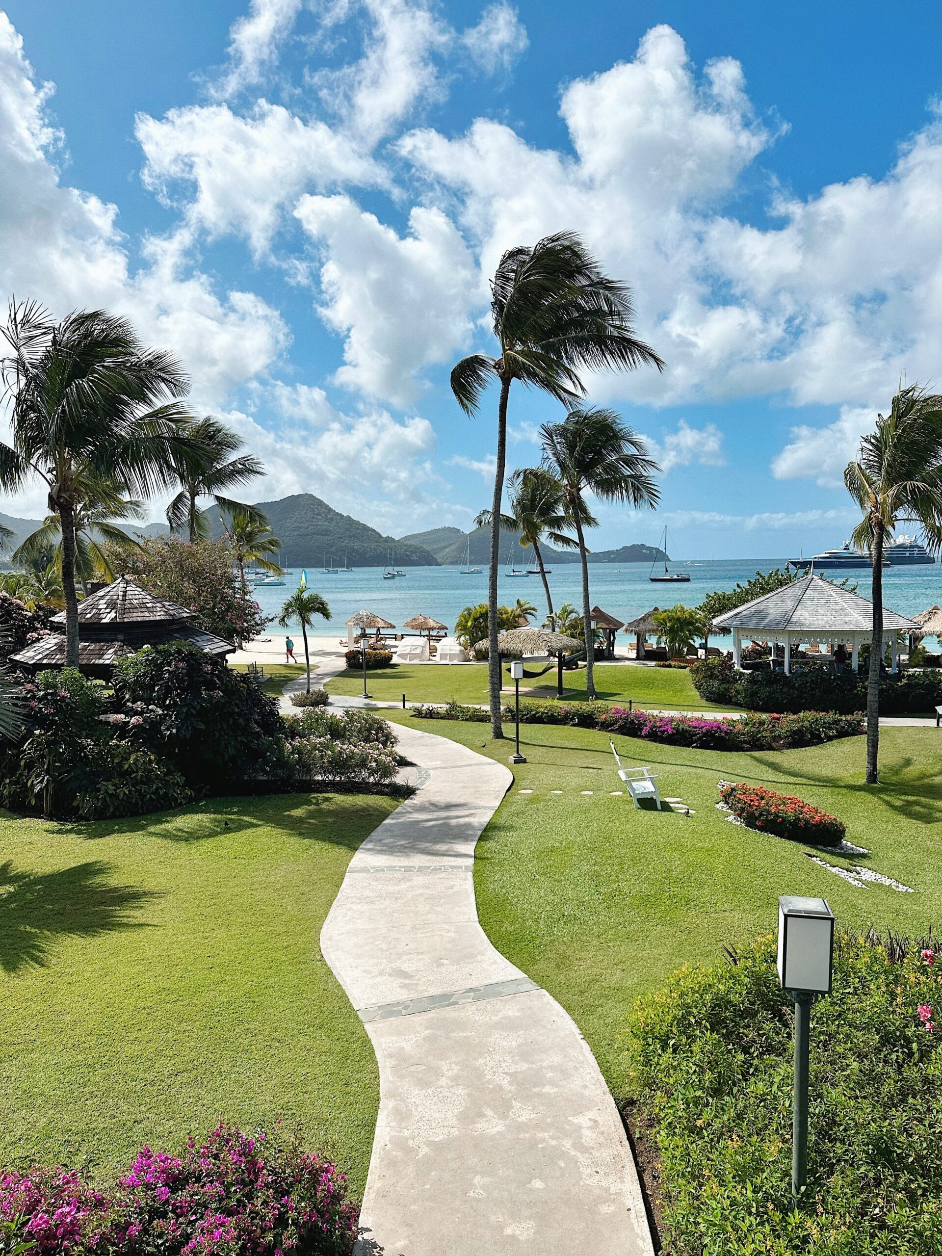 A serene tropical landscape features a winding path through lush greenery, palm trees, and colorful flowers, leading toward a beach with calm waters and distant mountains under a partly cloudy sky.